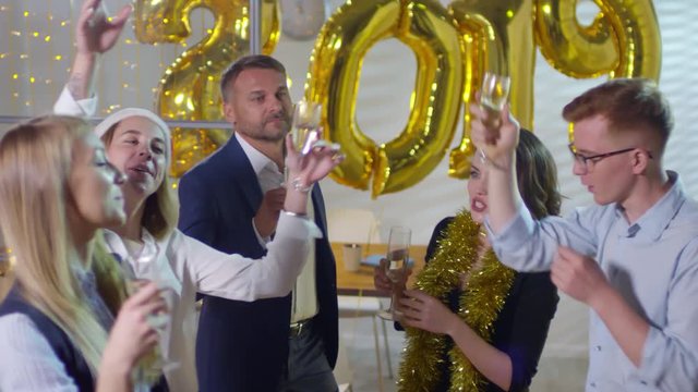 Handheld shot of happy businessmen and businesswomen holding glasses of champagne and dancing at New Years party in office with golden number 2019 balloons and Christmas lights