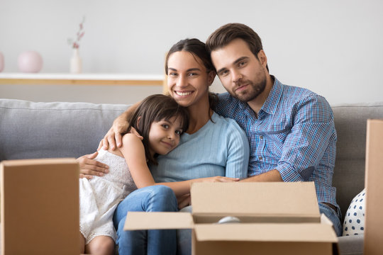 Happy diverse family sitting on couch in living room with carton boxes at move day feels happy and proud. Married couple and little daughter rest looking at camera. Buy new home and relocate concept