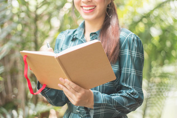 Happy Young Asian Woman Writing in Notebook and Smiling in Spring Garden.Asian women relax in the holiday. Study read a book. Read a book In the garden.