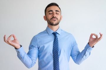 Calm handsome businessman with closed eyes meditating. Serene young employee in shirt and tie holding hands in mudra and focusing on thoughts. Relaxation concept
