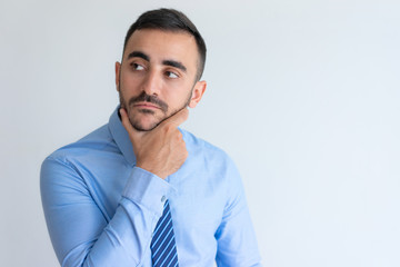 Pensive handsome young male manager in blue shirt touching chin and looking away. Confident businessman contemplating. Dilemma concept