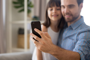 Smiling father and little cute preschool daughter looking at smartphone screen taking selfie portrait photography sitting on couch, focus on male hand close up. Family have fun use technology concept