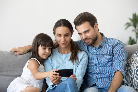 Married diverse couple spend free time with little daughter sitting together on couch in living room at home. Positive mother father small child using mobile phone watching cartoons play games online