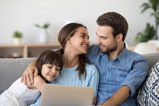 Multi-ethnic young married couple and little adorable daughter sitting on sofa at home spend free time on lazy weekend using pc watching interesting program funny videos laughing feels happy satisfied