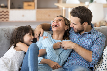 Cheerful people sitting on couch in living room have fun little daughter tickling mother laughing...