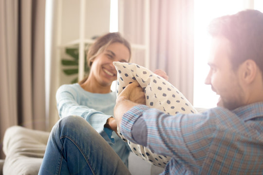 Cheerful Diverse Married Couple In Love Sitting On Sofa In Living Room At Home. Millennial Pretty Mulatto Wife And Caucasian Husband Have Fun Playing Fighting With Pillows Laughing Enjoying Free Time