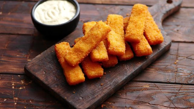 Pile of golden fried fish fingers with white garlic sauce placed on chopping board on dark wooden background.