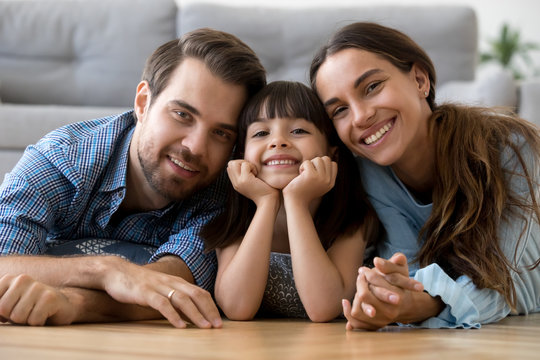 Multi-ethnic diverse family lying at cushions on warm floor in living room at modern home smiling looking at camera. Young mother adorable daughter handsome father feels happy spend free time together