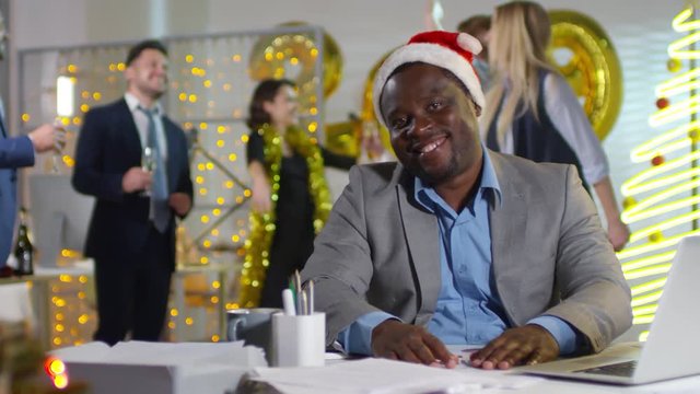 PAN of happy black office worker in Santa hat sitting at his desk and smiling for camera as his colleagues drinking champagne and dancing at New Years party at work. Number 2019 balloons in background