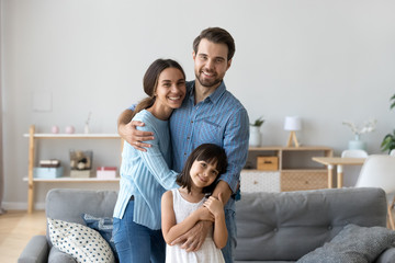 Cheerful diverse multi-ethnic family married couple wife husband little daughter embracing standing together in living room smiling looking at camera at new modern home feels happy and satisfied.