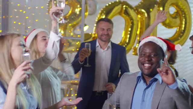 Handheld shot of happy black businessman in suit and Santa hat holding glass of champagne and dancing with colleagues during New Years office party. Golden number 2019 balloons in background