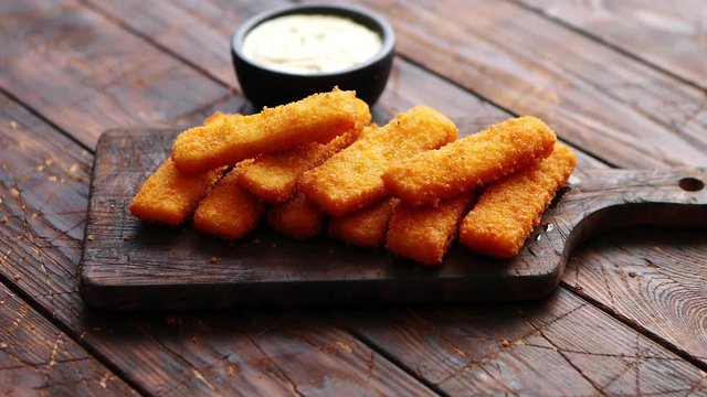 Pile of golden fried fish fingers with white garlic sauce placed on chopping board on dark wooden background.