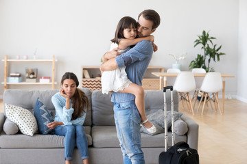 Unhappy diverse family in living room at home. Sad father hugs hold on hand small daughter, mother sitting on sofa. Parents divorcing, child stay with mommy dad leave with suitcase. Break up concept - Powered by Adobe