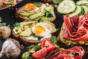 Various of sandwiches and bruschetta with prosciutto, fried quail egg, avocado, cucumber, tomatoes, spices on stone background