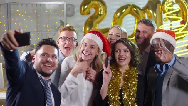 Handheld shot of happy businesspeople wearing tinsel and Santa hats posing for selfie at New Years party in office. Number 2019 golden balloons in background