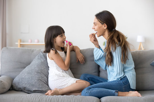 Funny little daughter with young mother or elder sister having fun together sitting on couch in living room at home looking at each other and smiling child copying female applying lipstick on lips.
