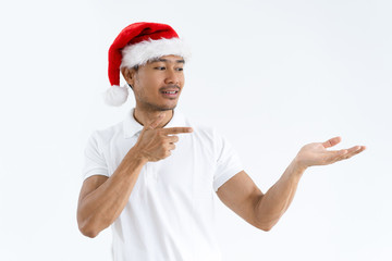 Fototapeta na wymiar Smiling Asian man wearing Santa Claus hat and presenting product. Guy holding empty space on palm and pointing at it. Christmas offer concept. Isolated front view on white background.