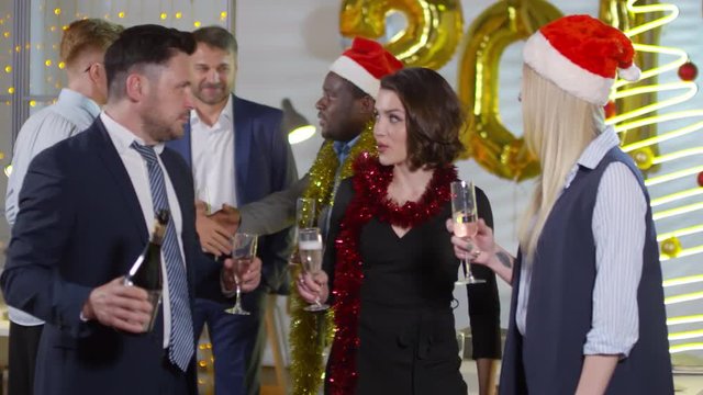 Medium shot of happy businessman in suit pouring champagne into glasses of happy female colleagues and toasting while having fun at office Christmas party