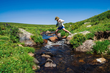 Naklejka na ściany i meble Traveler with camera on stone in mountain creek. Adventure of tourist. Hiking in mountains. Rich vegetation of highlands. Stream of clear water in brook. Vivid sunny landscape of majestic nature.