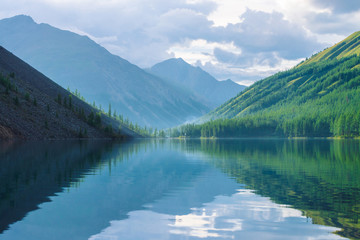 Naklejka na ściany i meble Ghostly mountain lake in highlands at early morning. Beautiful misty mountains reflected in calm clear water surface. Smoke of campfires. Amazing atmospheric foggy landscape of majestic nature.