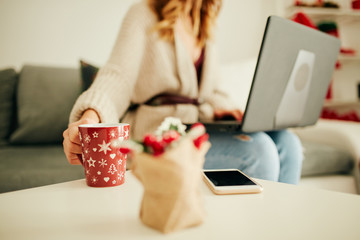 Woman holding cup of coffee while working on laptop at home for Christmas. New Year and Xmas concept.