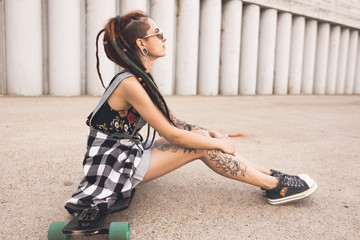 young girl with tattoo and dreadlocks sitting on a longboard on the background of concrete structure