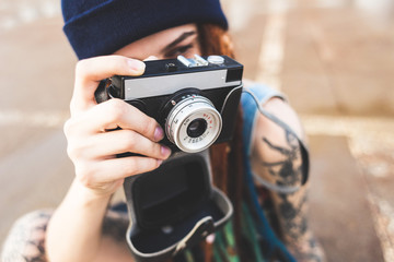 young girl with tattoos and dreadlocks in a blue hat photographs a vintage camera on the background of a concrete wall