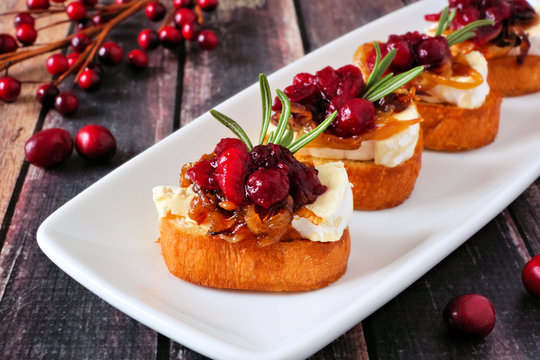 Crostini appetizers with cranberries, brie and caramelized onions. Close up on a serving plate against a wood background.