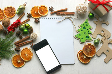 Fototapeta premium Christmas card, blank sheet of paper and mobile phone surrounded by Christmas balls, fir branches, cinnamon sticks, spices, dried orange, wooden deer 