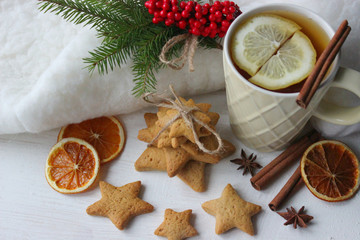 A Cup of tea with lemon on the table close-up surrounded by Christmas decorations and homemade cakes. Star shaped gingerbread, cinnamon sticks and dried oranges on white background.