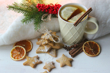 Obraz na płótnie Canvas A Cup of tea with lemon on the table close-up surrounded by Christmas decorations and homemade cakes. Star shaped gingerbread, cinnamon sticks and dried oranges on white background.