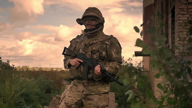 Strong authentical soldier is standing resistantly wearing camouflage and helmet with automatic gun in hands, freen trees and grass as background, empty space