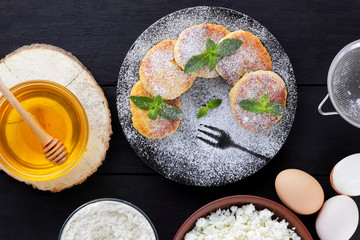 Cottage cheese pancakes and ingredients for their preparation on dark background. Syrniki with honey and mint on plate. Ingredients for cooking: flour, cottage cheese, eggs, sour cream, honey