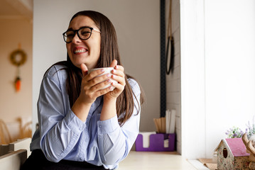 A woman with dark hair, in a blue shirt with coffee in her hands smiles very happily