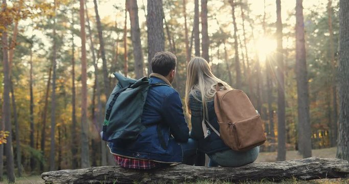 Back biew on the Caucasian young couple of hikers with big backpacks sitting and resting on the overthrown trunk in the wood and talking. Rear. At fall. Outside.