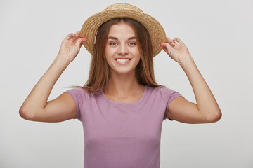 Cheerful young female with pleasant smile, has long brown hair, keeps in hands straw hat, tries it on or corrects, imagines a vacation by the sea, hot summer, against white background