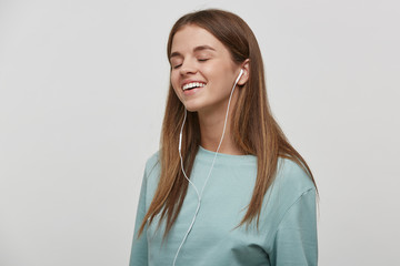 Obraz premium Keen on music young girl, smiles, listens favorite music with headphones, stands sideways, with closed eyes, dressed casual, on a white background in the studio.