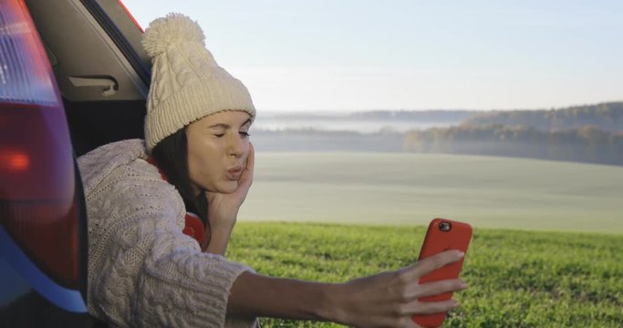 Young woman admiring the sunrise in trunk of car. Girl dressed in woolen socks taking photo on mobile phone against backdrop of forest panorama in fog . View from inside 4k video