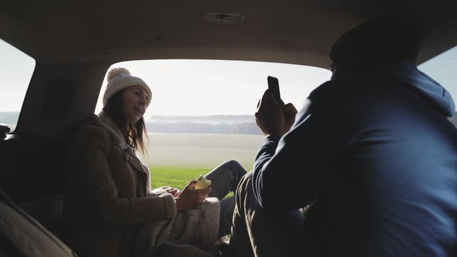 Personal perspective of couple lying in car's trunk, mountain landscape and admiring sunrise . Lovers alking and drinking hot coffee against backdrop of forest panorama in fog . View from inside