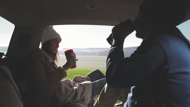 Personal perspective of couple lying in car's trunk, mountain landscape and admiring sunrise . Lovers alking and drinking hot coffee against backdrop of forest panorama in fog . View from inside