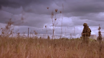 Resistant strong soldier holding automatic weapon and crossing wheat field alone in camouflage, dark cloudy sky during daytime, authentic view