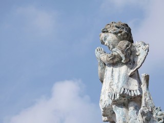 Stone statue of a little angel with wings, praying. Statue damaged by time. In the background, the...