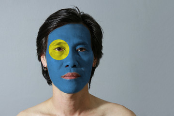 Portrait of a man with the flag of the Palau painted on his face on grey background. A light blue field with the large yellow disk shifted slightly to the hoist-side.