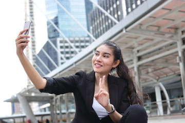 Attractive young Asian woman taking a photo or selfie with mobile smart phone on street of modern city.