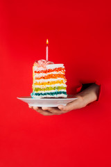 Piece of vegan rainbow birthday cake in woman's hand through torn red paper, selective focus