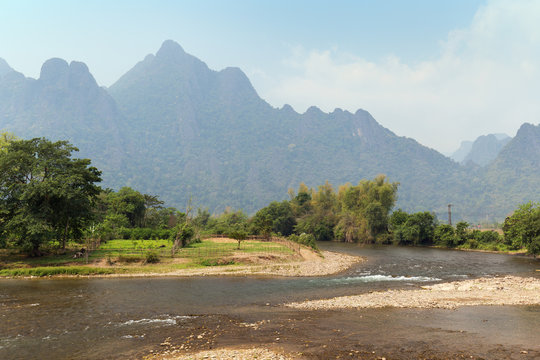 Scenic view of the Nam Song River at low tide, pasture and limestone mountains near Vang Vieng, Vientiane Province, Laos, on a sunny day.