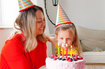 Adorable four years old european blonde girl makes a wish before blowing out the candles on a birthday cake that mom is holding