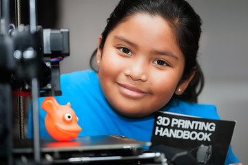 Cute girl smiling and showing her 3d printed creation on the bed of a 3d printer while holding a...