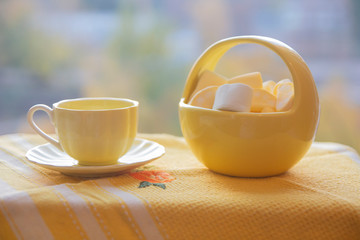  yellow coffee cup and ceramic yellow basket with marshmallows on a yellow tablecloth against the...