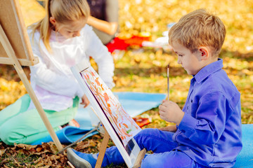 Cute little blond child boy in blue shirt drawing outdoor with other childern in autumn park....